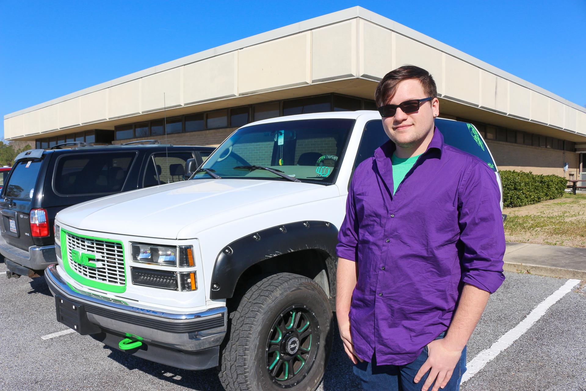 A man in a purple shirt stands in front of a white SUV.
