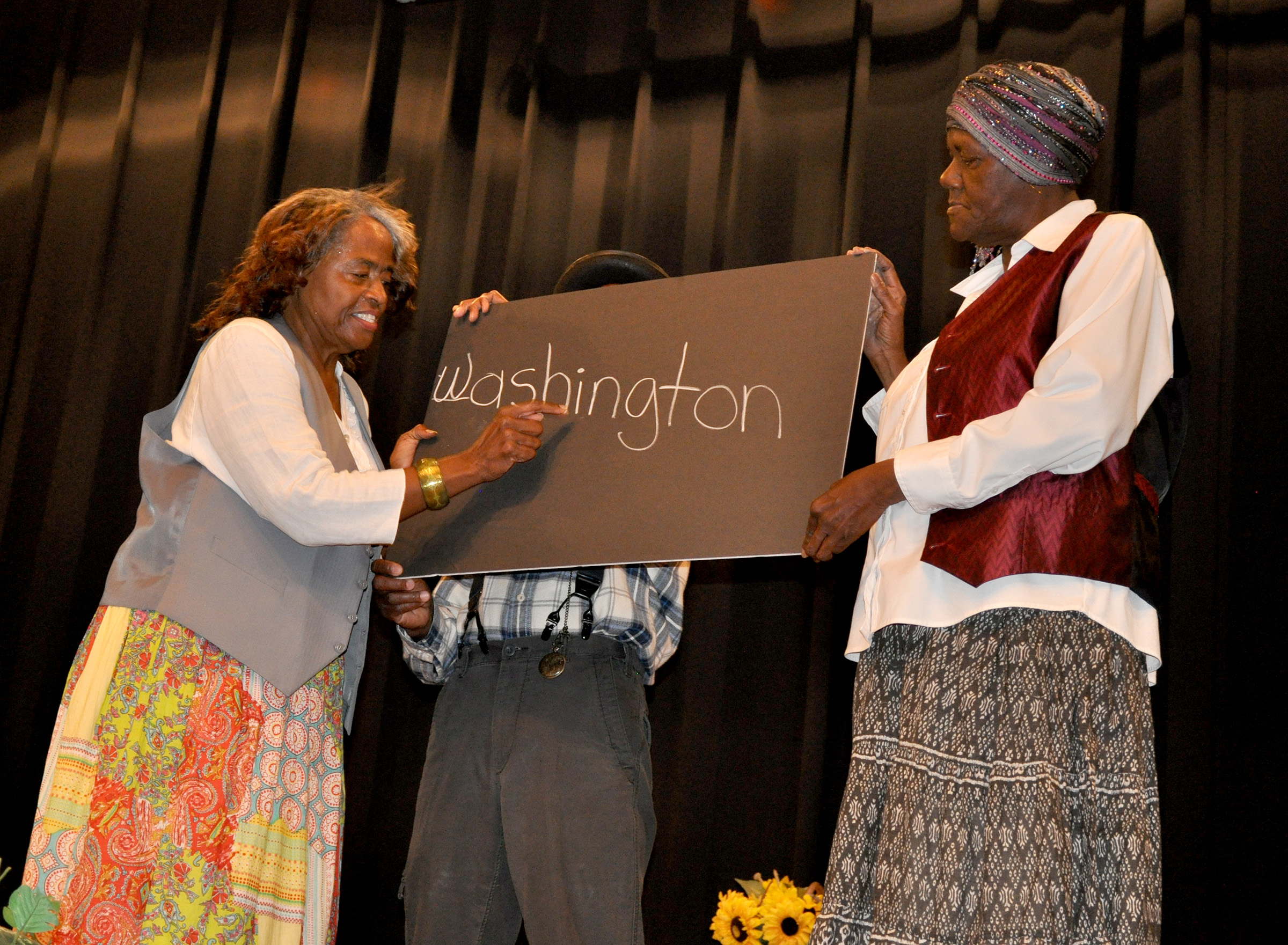 A woman writes the word Washington on a blackboard with two people holding the board.